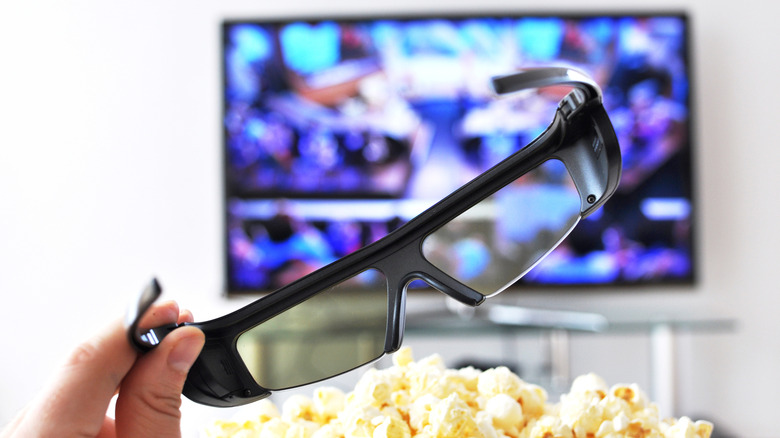 Active shutter 3D glasses resting on a table next to popcorn with a TV in the background.
