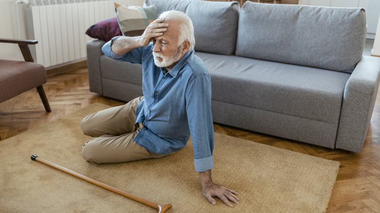 An elderly man lying on the floor after a fall, reaching for his head with a cane nearby.